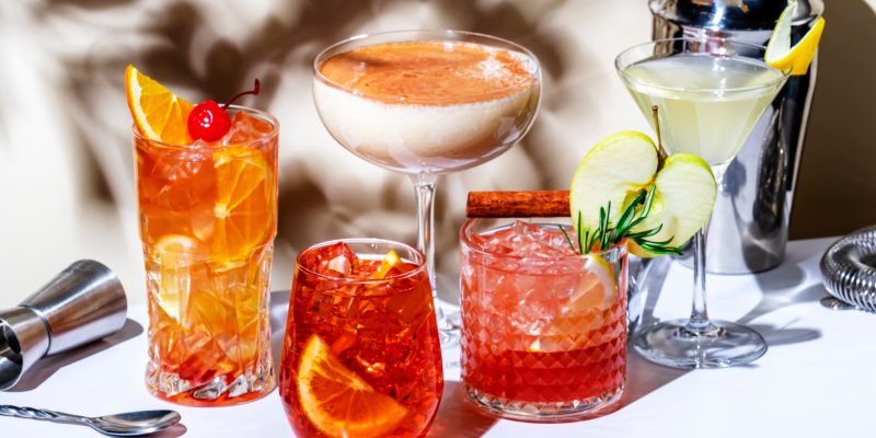 Fünf farbige Cocktails stehen auf einem Tisch, dekoriert mit Früchten und Gewürzen.