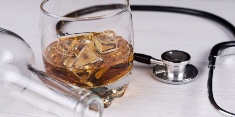 Glas mit Whiskey und Eiswürfeln, steht neben Stethoskop, auf weißem Tisch liegend.