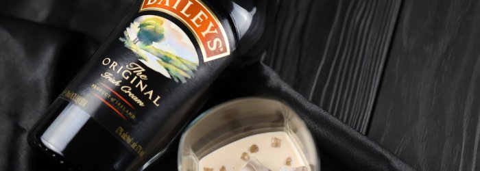 Bottle of Baileys lies next to filled glass with ice cubes on black fabric and table.
