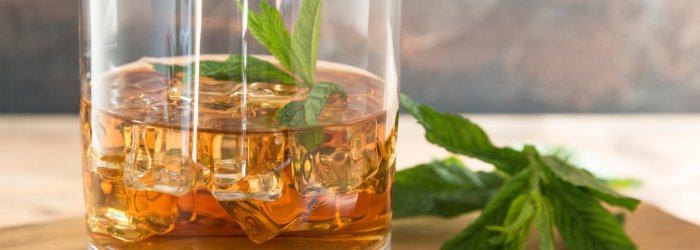 Verre contenant du whisky et des glaçons, décoré de feuilles de menthe, posé sur un support en bois.