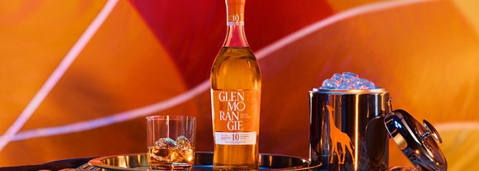 Bouteille de whisky avec verre et seau à glace sur un plateau, devant un fond coloré à motifs.