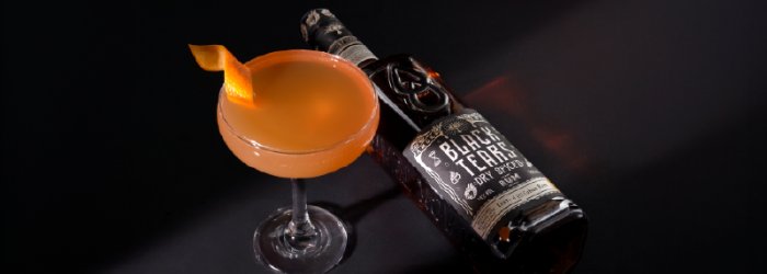 Ein oranger Cocktail im Glas neben einer Black Tears Flasche mit schwarzen Hintergrund