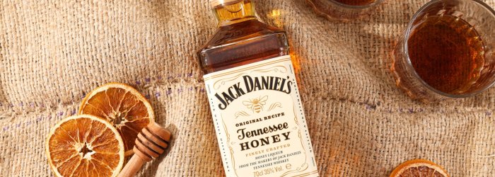 Bottle of Jack Daniel's Tennessee Honey lies on jute fabric, surrounded by glasses, cinnamon sticks and orange slices.