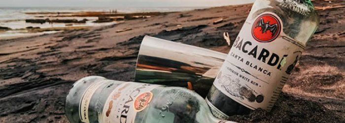 Zwei Bacardi-Flaschen liegen im Sand an einem Strand, Himmel bewölkt, Meer im Hintergrund.