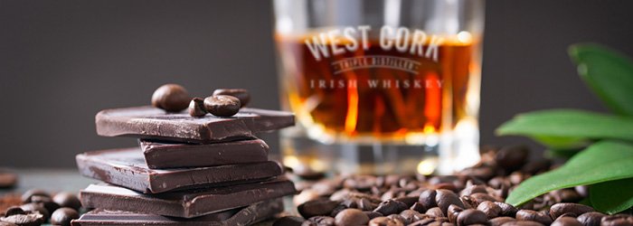 Un gros plan montre un arrangement raffiné d'éléments culinaires sur un fond sombre et feutré qui souligne la richesse des motifs. Au centre, un verre clair et court est rempli d'un liquide brun doré.