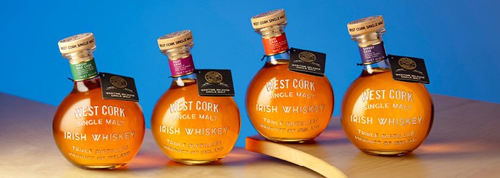 Four round bottles filled with a honey-colored liquid stand on a reflective surface against a graduated blue background. Each bottle is sealed with a cork and has a small black label on the neck