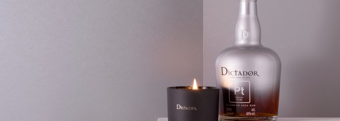Rum bottle next to a burning candle on a marble surface against a gray background. Text: "Dictador".