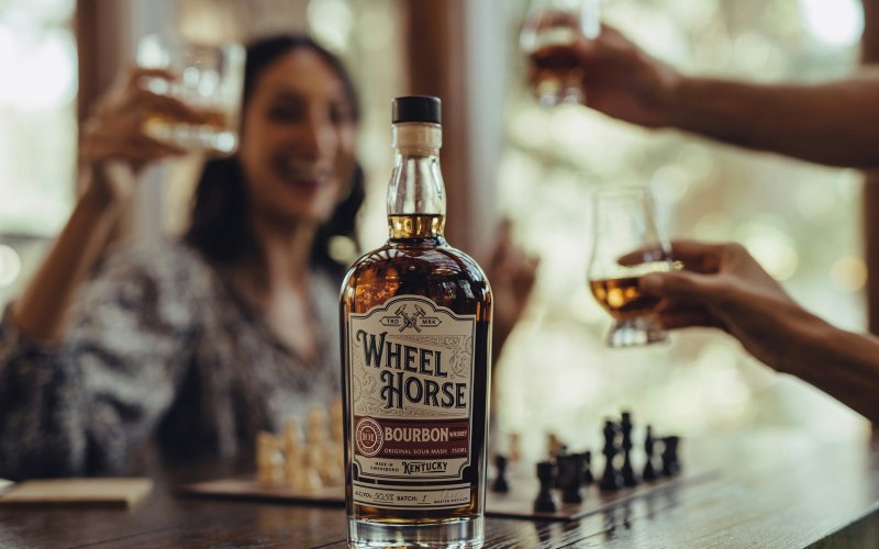 Bottle of bourbon "Wheel Horse" stands on wooden table while people clink glasses in the background.