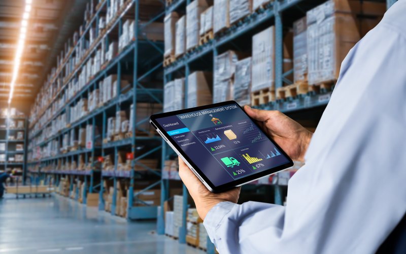 Tablette montrant la gestion des stocks, tenue par une personne en chemise bleue, devant des étagères de stockage bien remplies.