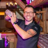 Un barman sourit et agite un shaker de cocktail dans un environnement de bar confortable.