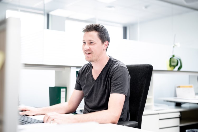 Een medium shot toont een geconcentreerde man met een lichte huid die ijverig aan het werk is achter een bureau in een modern kantoor, met een onscherpe achtergrond die zijn concentratie benadrukt. De man met kort donker haar en een lange